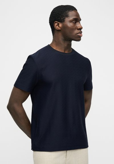 Άνδρας με κοντά σγουρά μαλλιά, φοράει μπλε σκούρο κοντομάνικο πουκάμισο και ανοιχτό μπεζ παντελόνι, στέκεται πλαγίως με τα χέρια πίσω από την πλάτη.