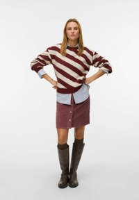 Strickpullover mit kastanienbraunen und cremefarbenen Streifen, über einem hellblauen Hemd mit Knöpfen getragen. Burgunderfarbener Cordrock mit Knöpfen und braunen kniehohen Stiefeln.