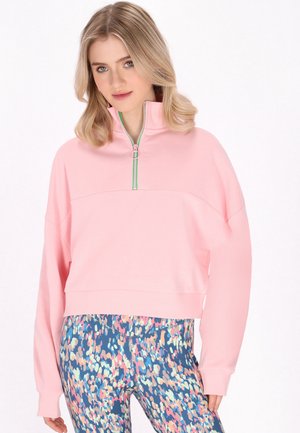 Ung kvinde med skulderlangt blondt hår bærer en lyserød sweatshirt med kvart-lynlås og leggings med flerfarvet abstrakt print.