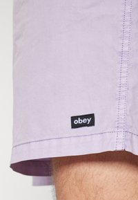 Světle fialové šortky vyrobené z hladké tkaniny. Mají černou štítkovou značku "obey" blízko lemu a viditelné detaily šití.