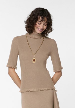 Vrouw met krullend haar, gekleed in een beige gebreide top met korte mouwen en een bijpassende rok, gecombineerd met een lange gouden ketting met een grote hanger.