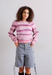 Gaiši rozā adīta džemperis ar purpura svītrām un ripotiem piedurknēm, apvienots ar pelēkiem denim šortiem ar sānu saitēm.
