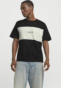 Camiseta de manga corta en negro y crema con un diseño horizontal y el logo "Originals Studio" centrado en la parte delantera. Material de algodón.
