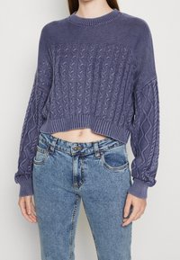 Dzianinowy fioletowy sweter cropped z wzorem w warkocze, z ribbowanym dekoltem i mankietami, w połączeniu z jasnoniebieskimi dżinsami o wysokim stanie.