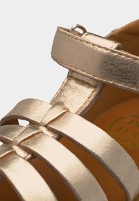 Κοντινή λήψη χρυσών μεταλλικών λουριών σανδαλιών με αγκράφα στο πάνω μέρος του ποδιού και υφή δερμάτινου υλικού.