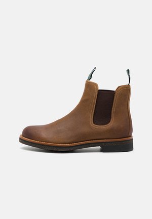 Bota Chelsea de cuero marrón con paneles elásticos en los laterales, punta redonda y tira verde en la parte superior. Cuenta con costuras en contraste y una suela de goma negra.