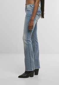 Moteris vilkinti šviesiai mėlynas bootcut džinsus ir juodas aukštakulnes aulinukes, stovinti šoninėje pozicijoje prieš vientisą baltą foną.