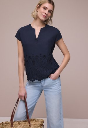 Giovane donna con camicetta ricamata blu navy e jeans azzurri tiene una borsa intrecciata, in piedi su sfondo beige semplice.