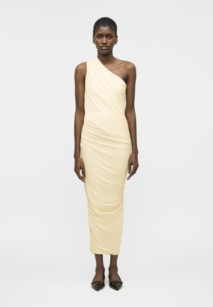 Kvinde iført en ærmeløs, enkeltskulder, flæset midi-kjole i cremefarve med sorte spidse flade sko, stående mod en ensfarvet baggrund.