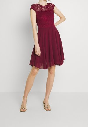 Woman wearing a knee-length sleeveless burgundy dress with lace bodice and chiffon skirt, paired with gold strappy high-heeled sandals.