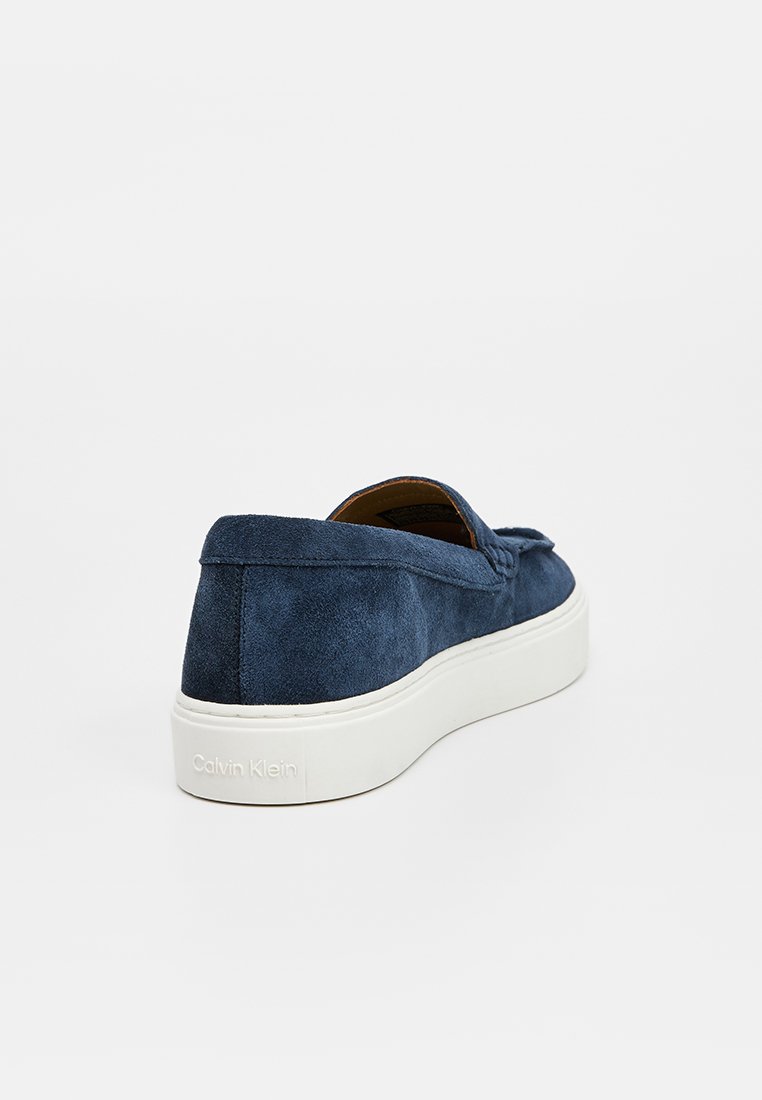 Chaussures slip-on en suède bleu marine avec une semelle blanche, présentant un design minimaliste et un détail logo subtil au niveau du talon.