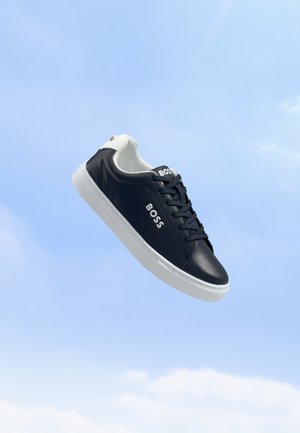 Schwarz-weißer BOSS-Sneaker mit weißer Sohle, der vor einem blauen Himmel mit leichten Wolken schwebt.