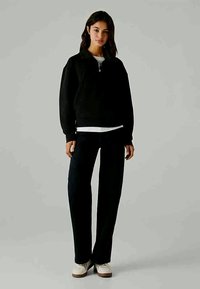 Čierna mikina na zips s rebrovanými manžetami, v kombinácii s tmavými džínmi. Model v béžových teniskách stojí proti neutrálnemu pozadiu.