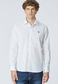 Giovane con capelli ricci indossa una camicia bianca con bottoni e un piccolo logo ricamato, abbinata a jeans blu, su uno sfondo semplice.