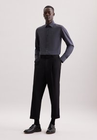 Camicia blu scuro con colletto e una sottile texture, abbinata a pantaloni neri a vita alta e scarpe nere lucide. Mani infilate nelle tasche.