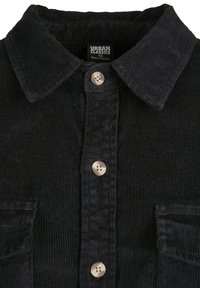 Chemise en velours côtelé noir avec col pointu, boutons sur le devant et deux poches poitrine, présentant une texture côtelée subtile. Les boutons sont de couleur claire.