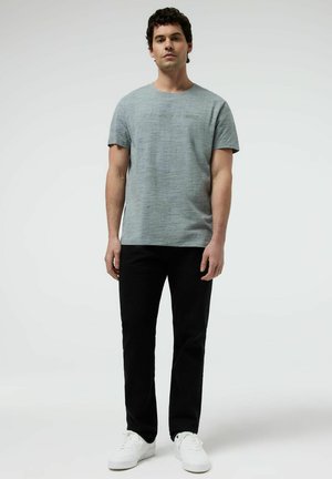 Mężczyzna stojący twarzą do przodu, ubrany w jasnoszarą koszulkę z krótkim rękawem, czarne spodnie i białe trampki na jednolitym tle.