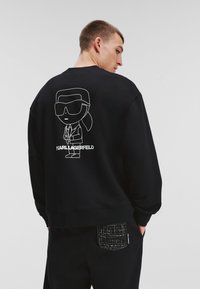 Sudadera negra con una figura de dibujo animado con gafas de sol en la parte trasera y el texto "KARL LAGERFELD" debajo. Tejido suave, corte relajado.