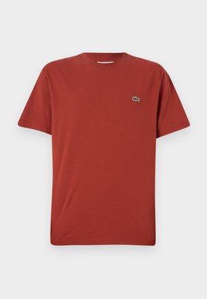 T-shirt in cotone rosso con colletto tondo e maniche corte, caratterizzato da un piccolo logo di coccodrillo verde sul lato sinistro del petto. Texture liscia.
