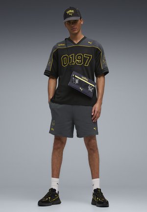Jonge man die een zwart-gele Pokémon x Puma outfit draagt, inclusief pet, mesh shirt, korte broek, schoudertas, sokken en sneakers, staand.