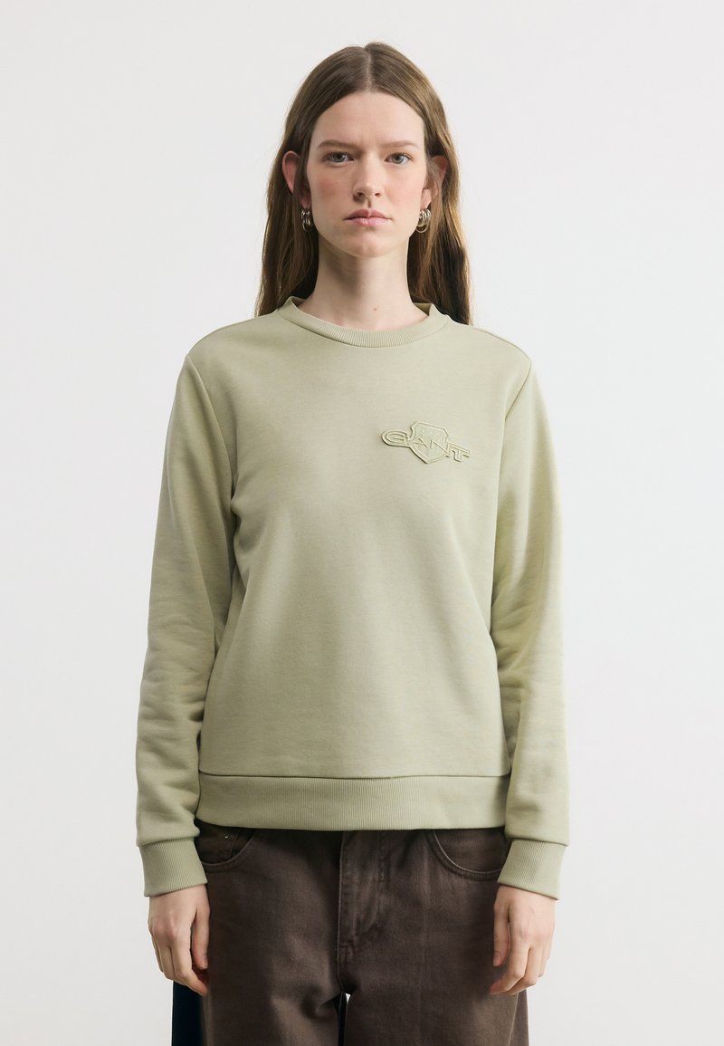 Γυναίκα με μακριά καστανά μαλλιά φοράει ανοιχτό πράσινο φούτερ και σκούρο καφέ παντελόνι, στέκεται μπροστά σε απλό, ανοιχτόχρωμο φόντο.