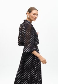 Svart prickig klänning med en genomskinlig, flödande design, breda ärmar och ett bälte i midjan. Kännetecknas av sina ljusgrå prickar och eleganta siluett.