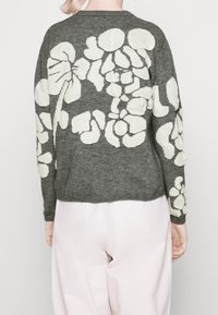 Personne portant un pull en maille gris foncé avec de grands motifs floraux blancs abstraits dans le dos et sur les manches, associé à un pantalon rose clair.