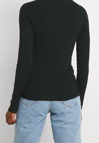 Haut à manches longues côtelé vert foncé avec col montant, associé à un jean en denim bleu clair avec poches arrière et coupe légèrement décontractée.