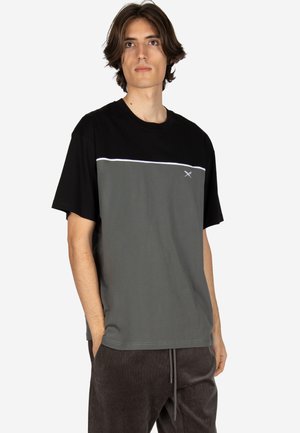 Junger Mann mit mittellangem Haar, der ein schwarz-graues kurzärmeliges T-Shirt und dunkle Hosen trägt, steht mit einer Hand in der Tasche.