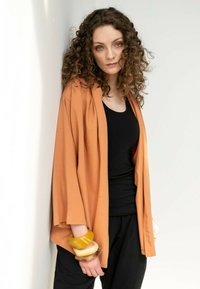 Długi rękaw, otwarty kardigan w kolorze terakoty, wykonany z miękkiego materiału. Noszony na czarnym topie na ramiączkach, uzupełniony kolorowymi bransoletkami.