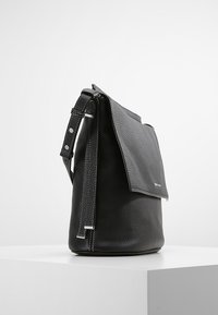 Borsa a tracolla in pelle nera con superficie texture, chiusura a pattina, tracolla regolabile e dettagli in metallo argentato. Forma cilindrica con base ampia.