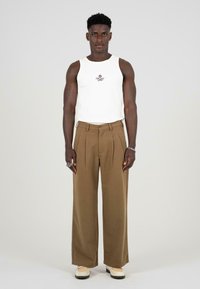Top sin mangas blanco con logo, combinado con pantalones de pierna ancha marrones. Los pantalones tienen un pliegue frontal y están hechos de una tela texturizada.