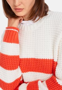 Strickpullover mit orange-weißen Streifen, strukturiertem Muster, gerippten Bündchen und rundem Ausschnitt. Nahaufnahme der Stoffdetails.