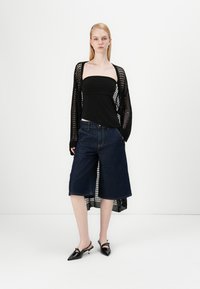 Cardigã em malha preta sobre um top preto sem alças, combinado com culotes de denim azul-escuro e sapatos pretos pontudos com tiras finas.