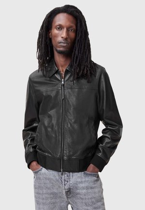 Giubbotto bomber in pelle nera con chiusura a zip, colletto e polsini a costine. Texture liscia e design aderente, abbinato a jeans grigio chiaro.