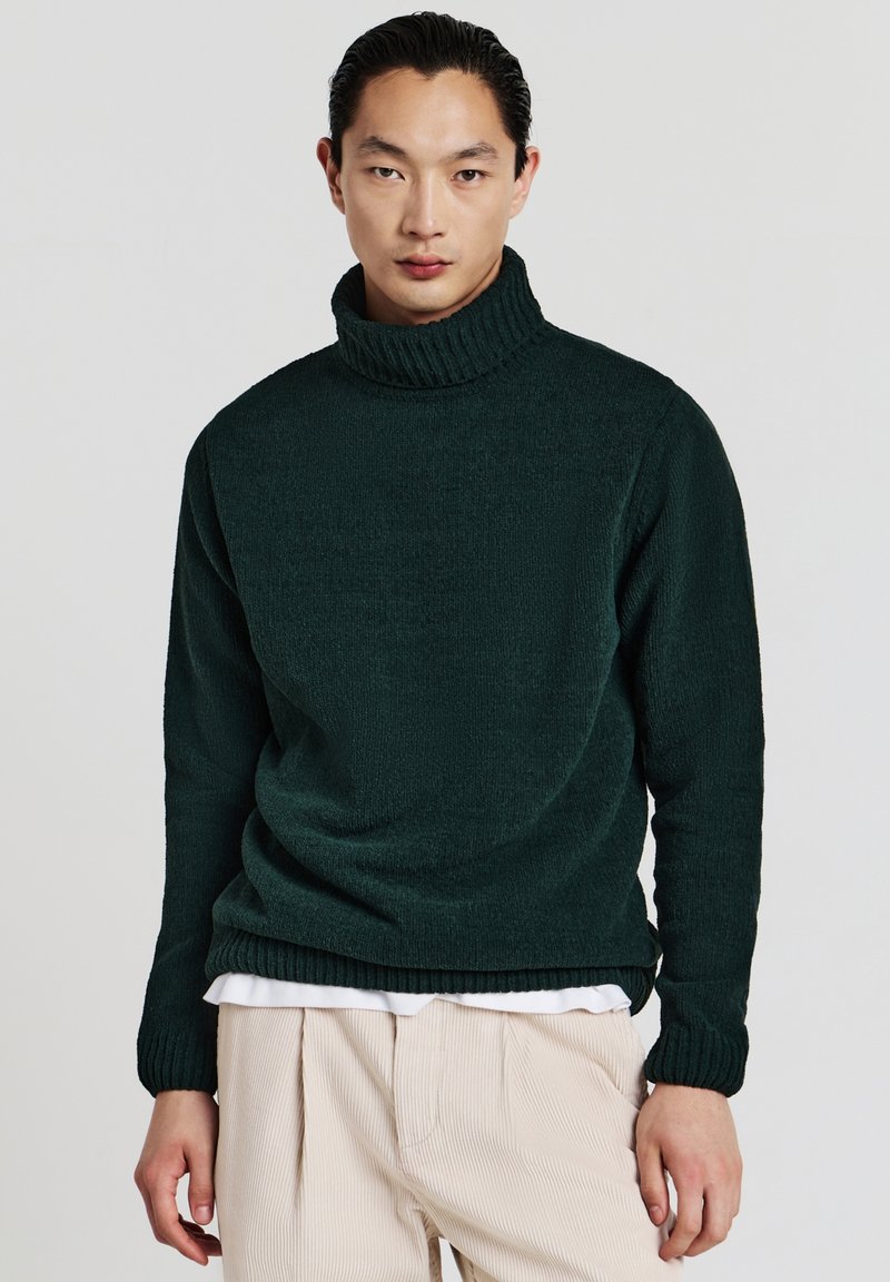 Dunkelgrüner Rollkragenpullover aus strukturiertem Stoff, mit hohem Kragen und gerippten Bündchen. Getragen über einem weißen Hemd und hellen Hosen.