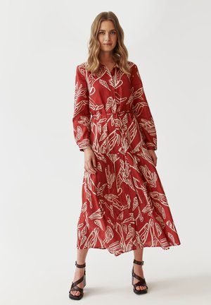 Rotes Hemdkleid mit einem weißen Blättermuster, tailliertem Schnitt, langen Ärmeln und einem gestuften Rock. Kombiniert mit dunklen Riemchen-Heels.