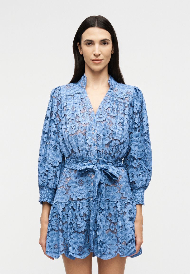 Woman with long dark hair wearing a blue floral lace dress with puffed sleeves and a tied waist belt, facing forward against a plain background.