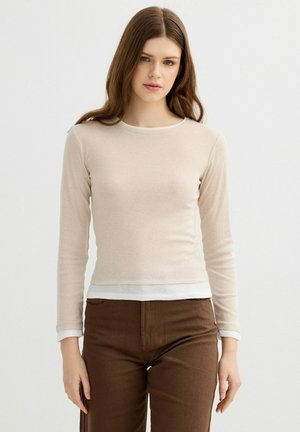 Mujer joven con cabello largo y castaño que lleva un top beige de manga larga sobre una camisa blanca y pantalones marrones, de pie frente a un fondo blanco.