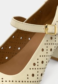 Μπεζ δερμάτινο παπούτσι Mary Jane με διάτρητο σχέδιο, χαμηλό τακούνι και λουράκι στον αστράγαλο με χρυσή αγκράφα. Ομαλή υφή και μινιμαλιστικό σχήμα.