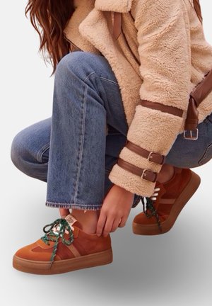 Personne en jean bleu et veste beige en peau lainée nouant des lacets verts sur des baskets marron tout en s'accroupissant sur un fond blanc.