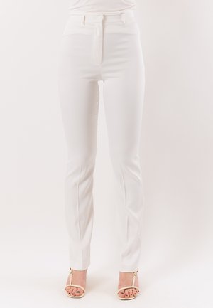 Pantaloni bianchi slim-fit con zip frontale e vita alta, indossati con tacchi aperti beige su uno sfondo chiaro uniforme.