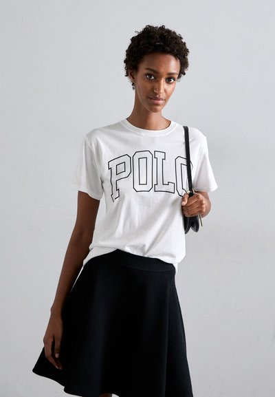 T-shirt en coton blanc avec "POLO" imprimé en grandes lettres noires, associé à une jupe noire, évasée et arrivant aux genoux. Design simple, look décontracté.
