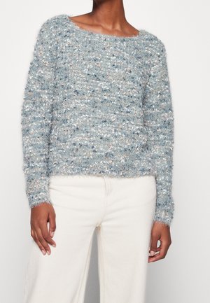 Femme portant un pull en maille texturé et duveteux bleu clair et gris à manches longues avec un pantalon blanc taille haute.