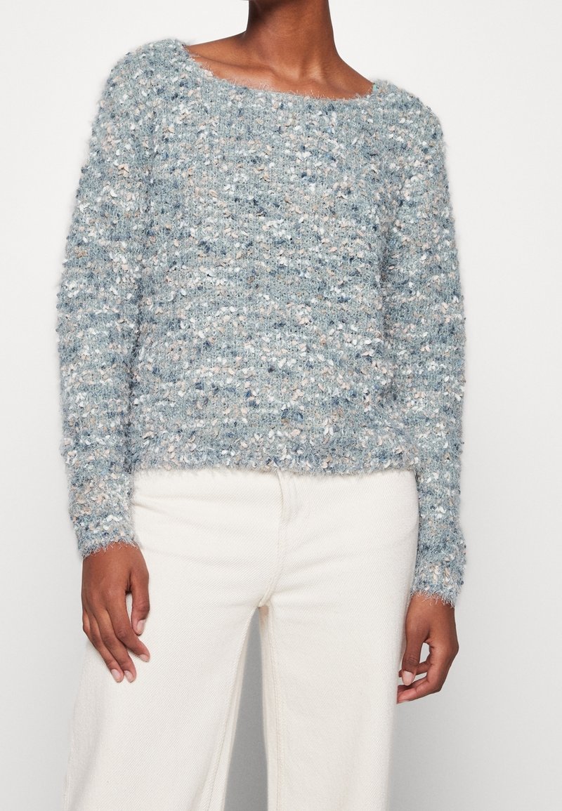 Femme portant un pull en maille texturé et duveteux bleu clair et gris à manches longues avec un pantalon blanc taille haute.