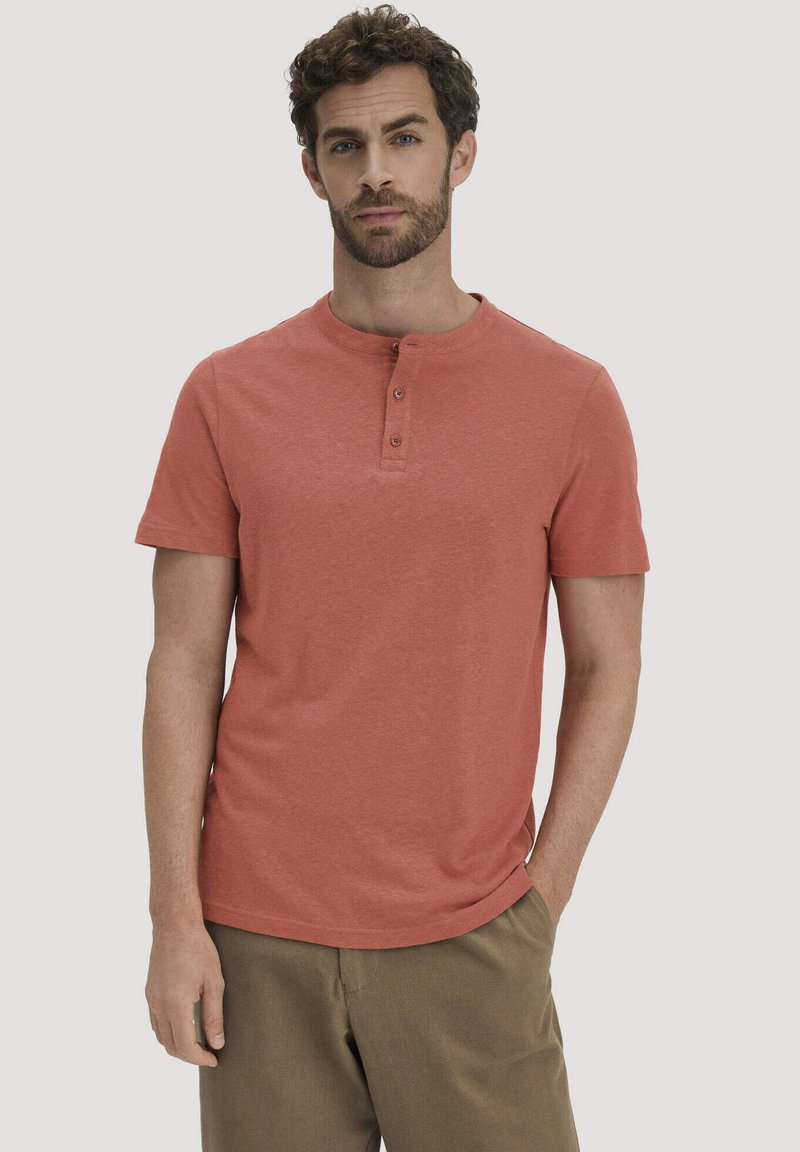 Man met krullend haar en baard, gekleed in een rustkleurig Henley-shirt met korte mouwen en een broek in bruintinten, staand met één hand in de zak.