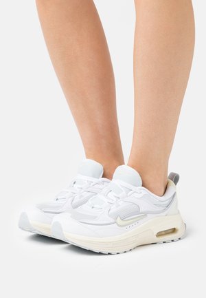Ben med vita Nike-sneakers som har mesh- och läderdetaljer, stående på en vit yta med en enkel bakgrund.