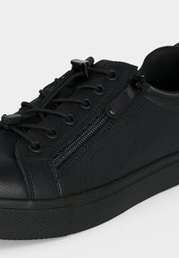 Baskets basses noires avec attaches élastiques pour lacets, fermeture éclair latérale, semelle texturée et dessus en cuir lisse.