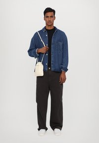 Giacca di denim blu sopra una maglietta nera, abbinata a pantaloni neri a gamba larga e sneakers bianche. Una piccola borsa a tracolla color crema è tenuta in una mano.