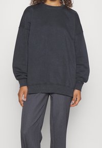 Sweatshirt oversized grigio scuro realizzato in tessuto morbido, con spalle scese e scollatura rotonda, abbinato a pantaloni grigi sartoriali.